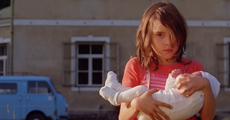 Filmstill aus "Die Vaterlosen": Ein Mädchen hält ein Baby im Arm und steht vor einem großen Haus. 